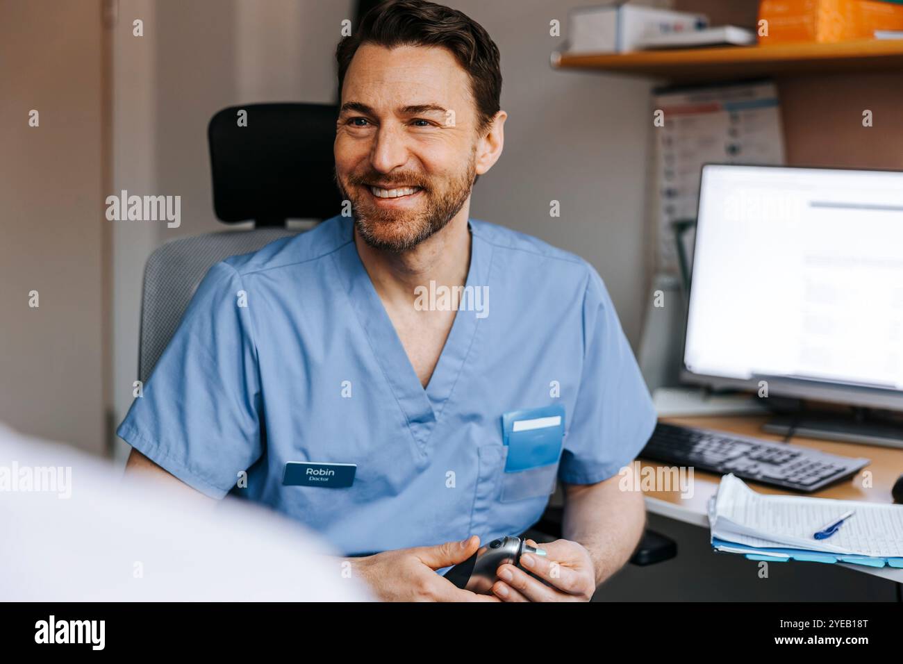 Felice esperto medico maschio che tiene il termometro auricolare mentre parla con il paziente in sala esame Foto Stock