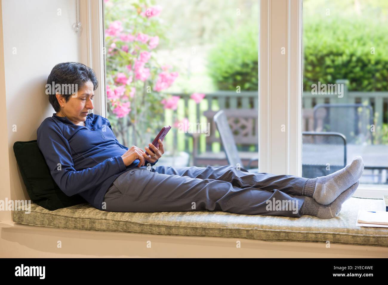 Donna asiatica indiana di mezza età che si rilassa seduta su un posto vicino alla finestra con i piedi in alto, guardando lo schermo del telefono con un giardino inglese sullo sfondo Foto Stock
