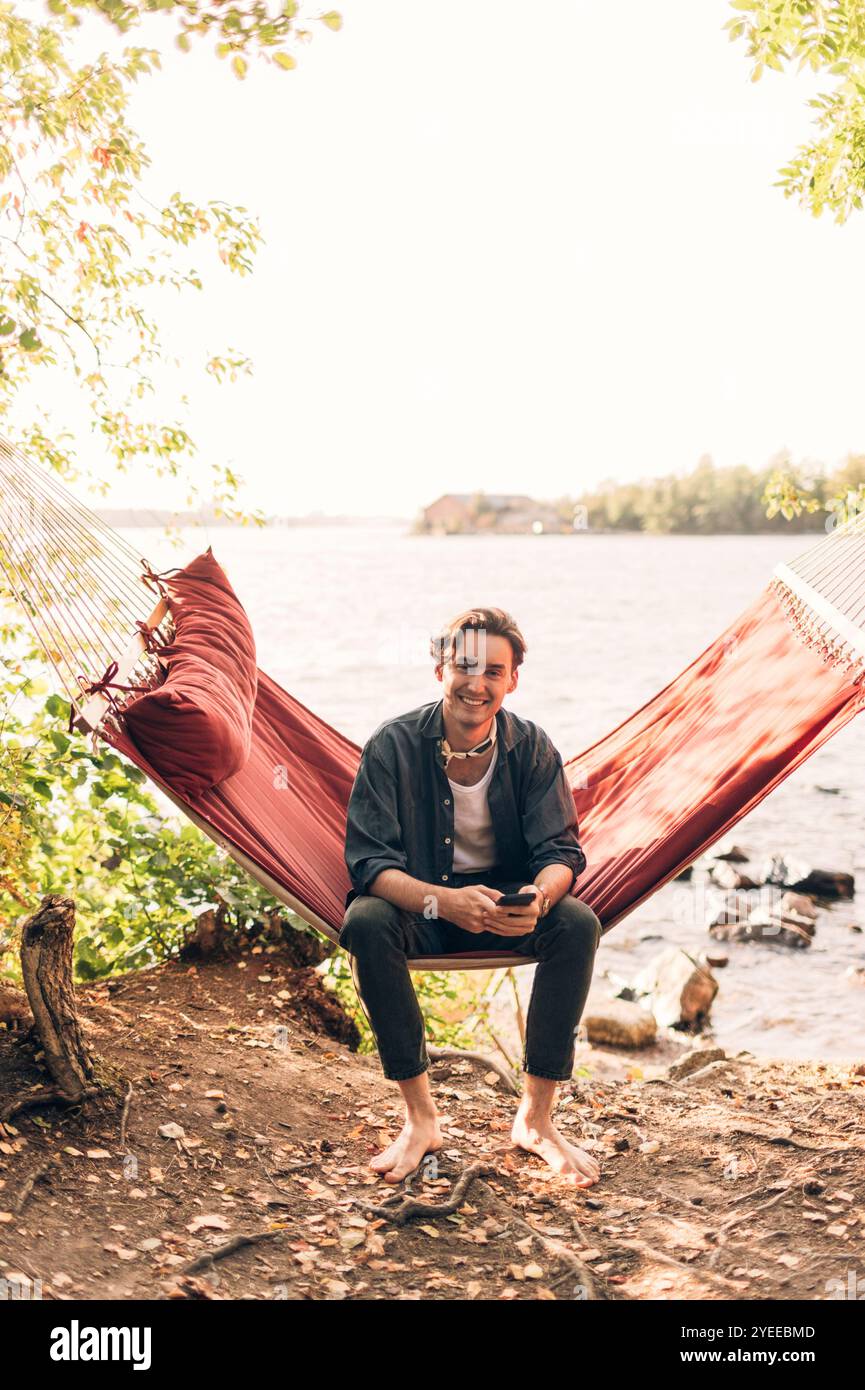 Ritratto di un uomo sorridente che tiene in mano uno smartphone mentre è seduto sull'amaca vicino al lago Foto Stock