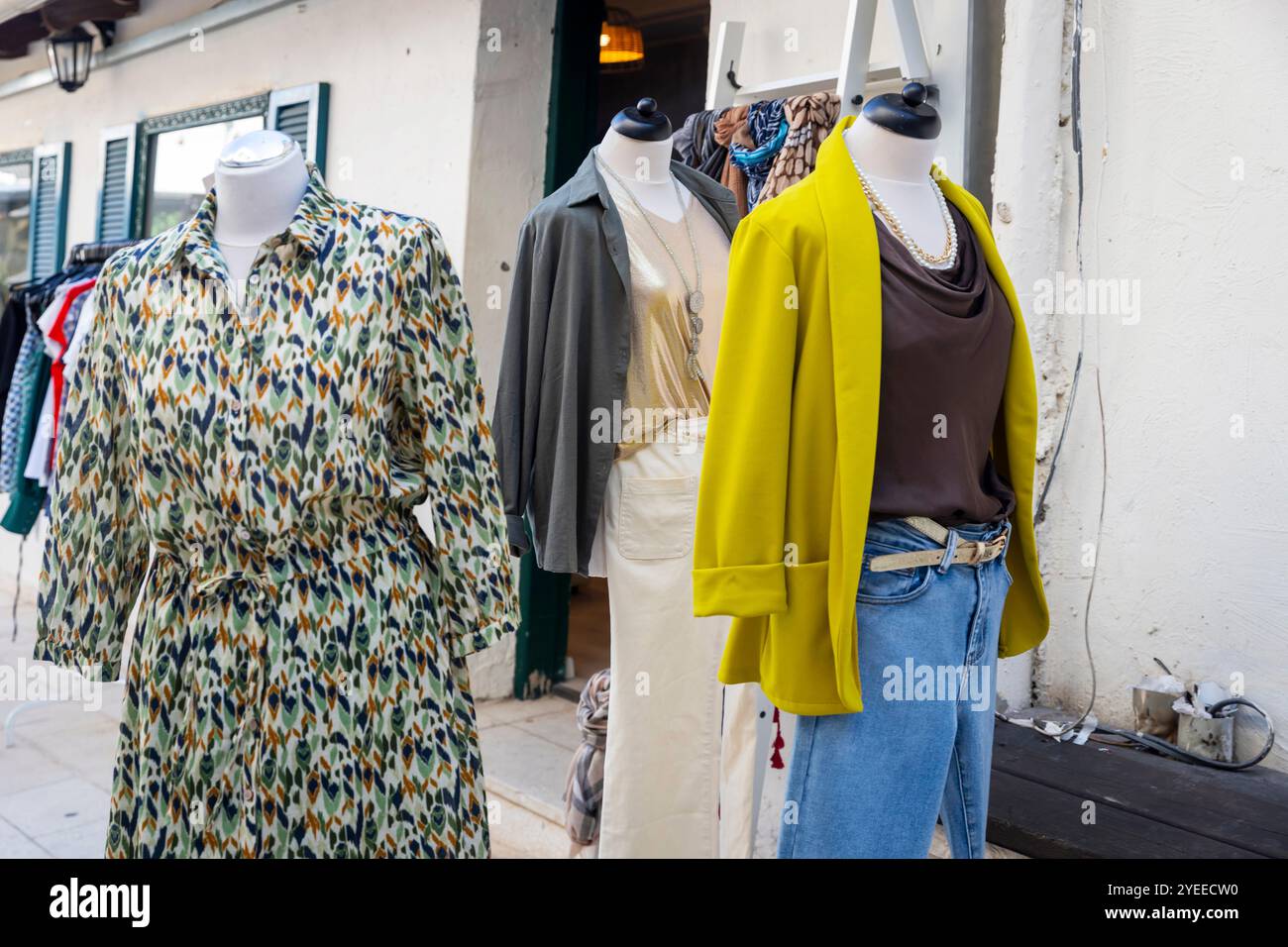 Manichini con abito verde-grigio, cardigan giallo, jeans blu e blazer grigio con pantaloni bianchi vicino a un negozio lungo la strada. Foto Stock