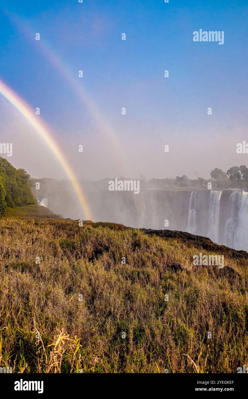 Un vibrante doppio arcobaleno si innalza sullo spruzzo nebbioso delle Cascate Vittoria, creando uno spettacolo naturale mozzafiato all'alba, durante la stagione secca. Foto Stock