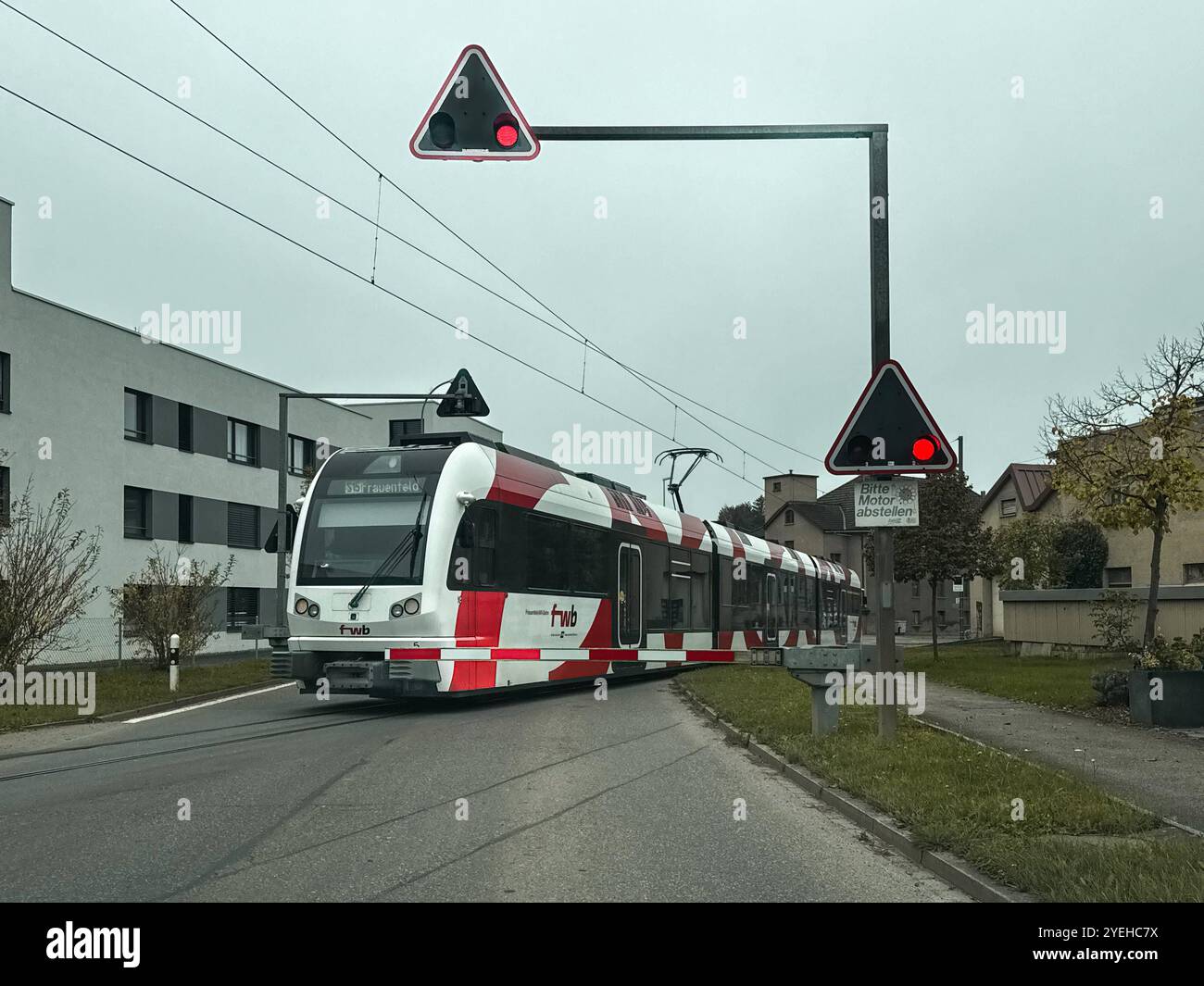 Frauenfeld, Schweiz, Europa, fwb-Zug, rote Ampel, Bahnübergang, Bitte Motor abstellen-Schild, Herbst, Verkehrssicherheit, CO2-Reduktion, Energieinsparung, ÖPNV, Emissionsvermeidung, Luftqualität, Energiekosten, Nachhaltigkeit, Stadtentwicklung, Klimaschutz, Infrastruktur, Mobilitätswende, Verkehrsplanung, Öffentlicher Nahverkehr, Schweiz, Signal, Schienenverkehr, Umweltbewusstsein, moderner Nahverkehr *** Frauenfeld, Svizzera, Europa, treno in direzione opposta, semaforo rosso, passaggio a livello, spegnere il segnale del motore, autunno, sicurezza stradale, riduzione di CO2, risparmio energetico, trasporto pubblico, emissioni avoi Foto Stock