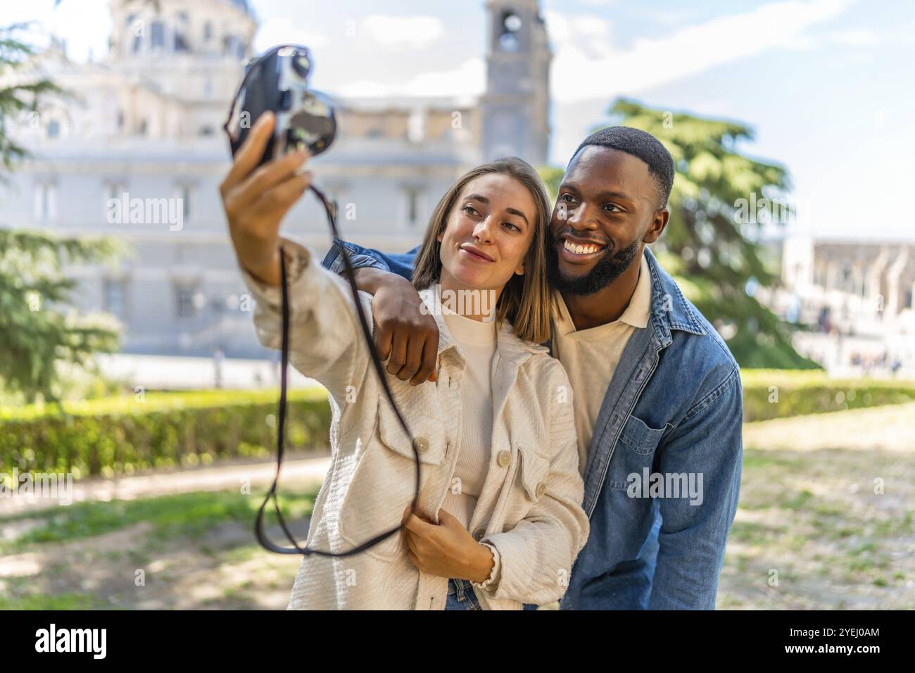 Felice coppia multietnica che scatta selfie con la fotocamera digitale in un parco con vista sul centro città Foto Stock
