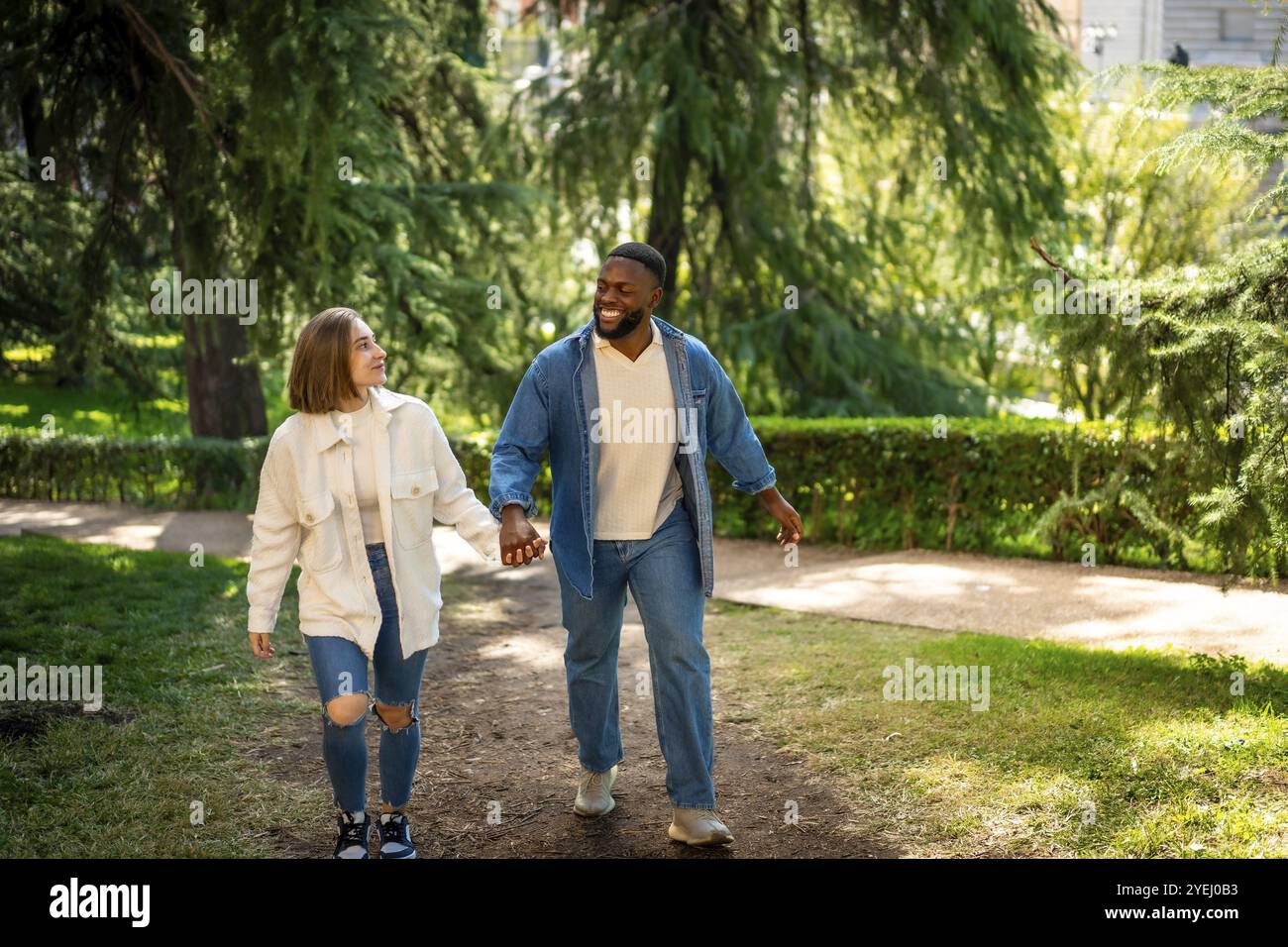 Felice coppia multietnica che indossa abiti casual in denim che camminano tenendosi per mano in un parco Foto Stock