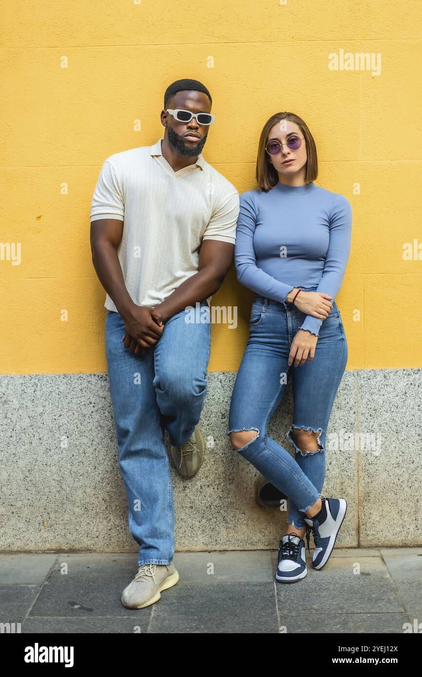 Coppia moderna e multietnica con abiti in denim e occhiali da sole alla moda appoggiati su pareti urbane Foto Stock