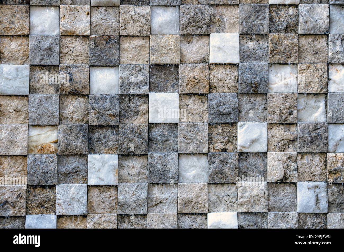 Parete composta da piccole piastrelle quadrate disposte a griglia. Le piastrelle variano per colore e consistenza, con sfumature di grigio, beige e bianco. Foto Stock