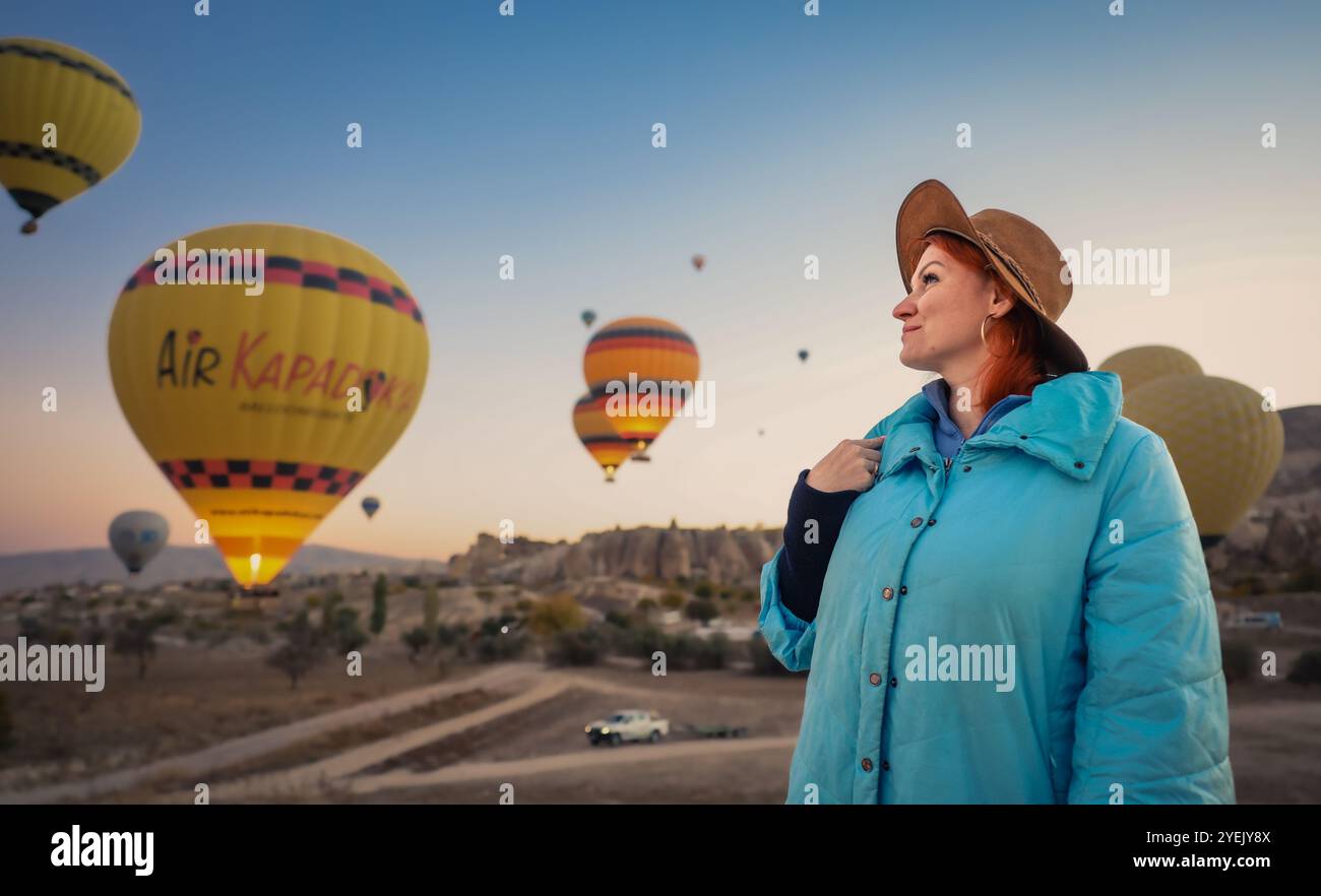 Mentre l'alba si apre in Cappadocia, una donna guarda con stupore le vivaci mongolfiere che si innalzano sopra aggiungendo un tocco magico alle formazioni rocciose uniche Foto Stock