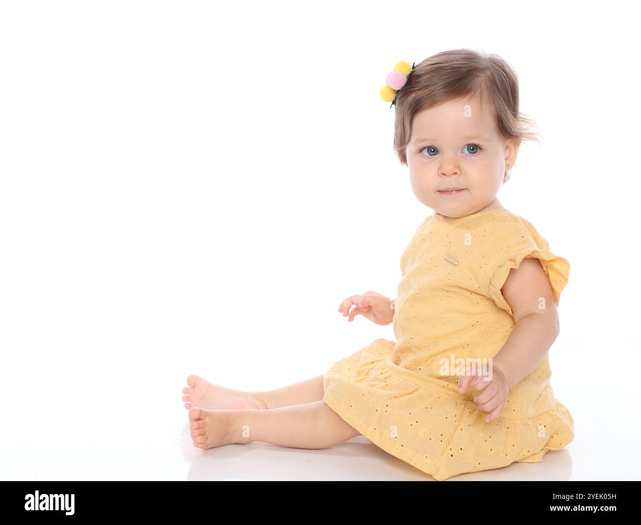 Bellissima Baby Girl dagli occhi blu in Studio Photo con indosso un abito giallo seduta su uno sfondo bianco, guardando verso la fotocamera. Foto Stock