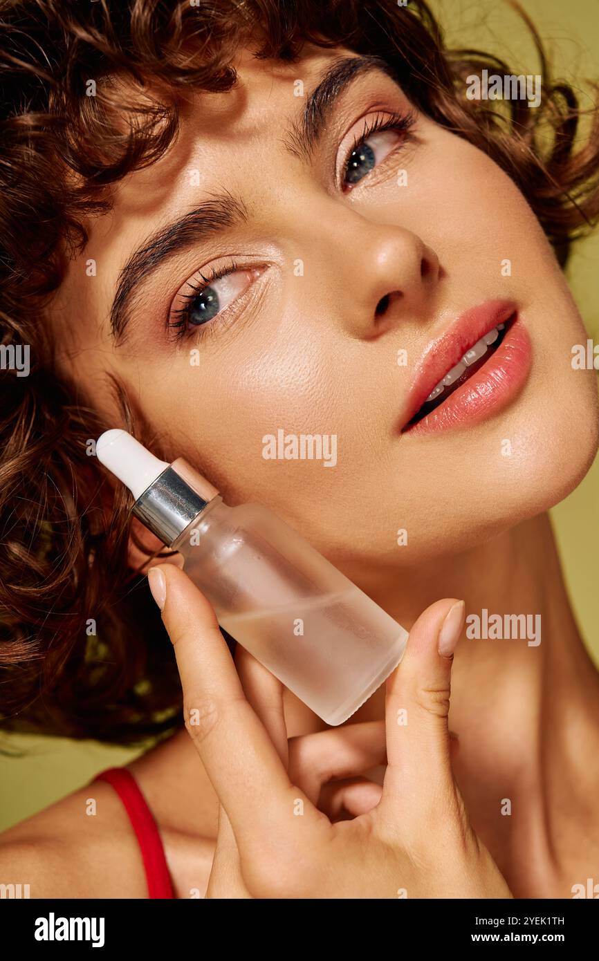Una giovane donna con capelli ricci applica teneramente il siero sulla pelle, irradiando bellezza e sicurezza. Foto Stock