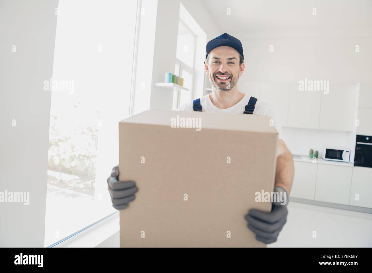 Tutore sorridente che consegna una grande scatola di cartone in un moderno interno della cucina, che mette in mostra un servizio di ristrutturazione professionale con facilità ed efficienza Foto Stock