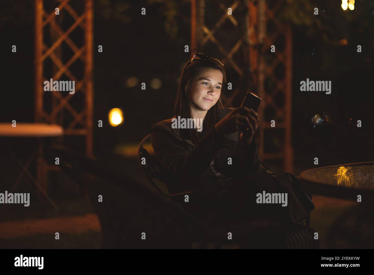 Una giovane donna sorridente che usa uno smartphone di notte. È seduta in giardino con una casa sullo sfondo Foto Stock