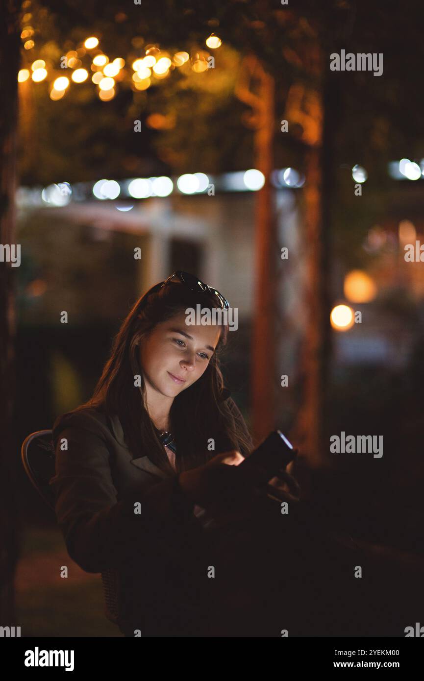 Una giovane donna sorridente che usa uno smartphone di notte. È seduta in giardino con una casa sullo sfondo Foto Stock