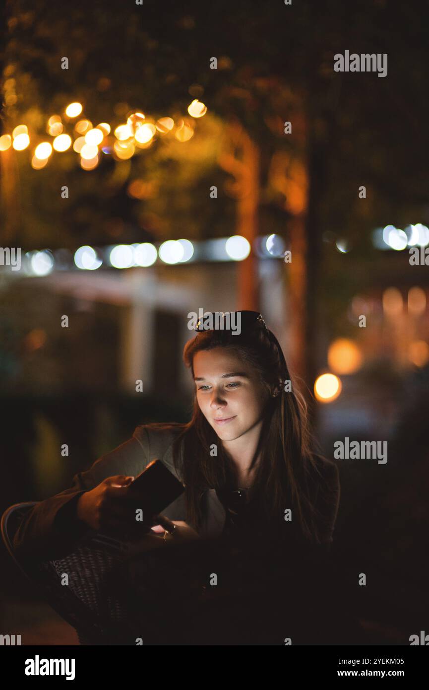 Una giovane donna sorridente che usa uno smartphone di notte. È seduta in giardino con una casa sullo sfondo Foto Stock