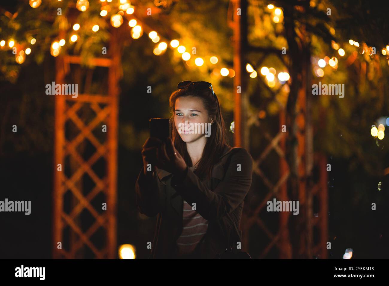 Una giovane donna sorridente che usa uno smartphone di notte. È seduta in giardino con una casa sullo sfondo Foto Stock