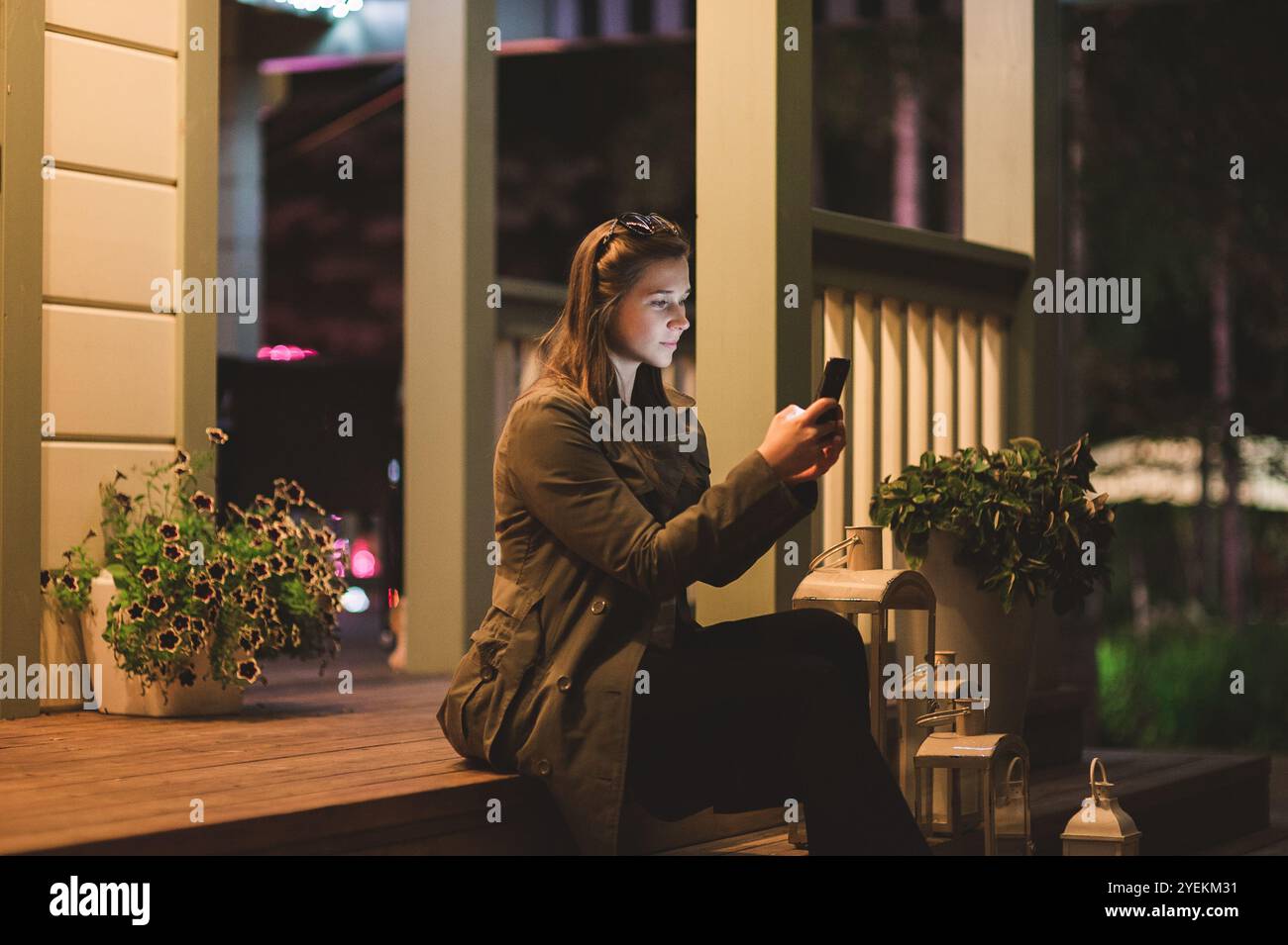 Una giovane donna sorridente che usa uno smartphone di notte. È seduta in giardino con una casa sullo sfondo Foto Stock