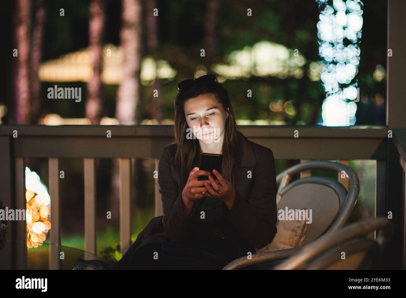 Una giovane donna sorridente che usa uno smartphone di notte. È seduta in giardino con una casa sullo sfondo Foto Stock