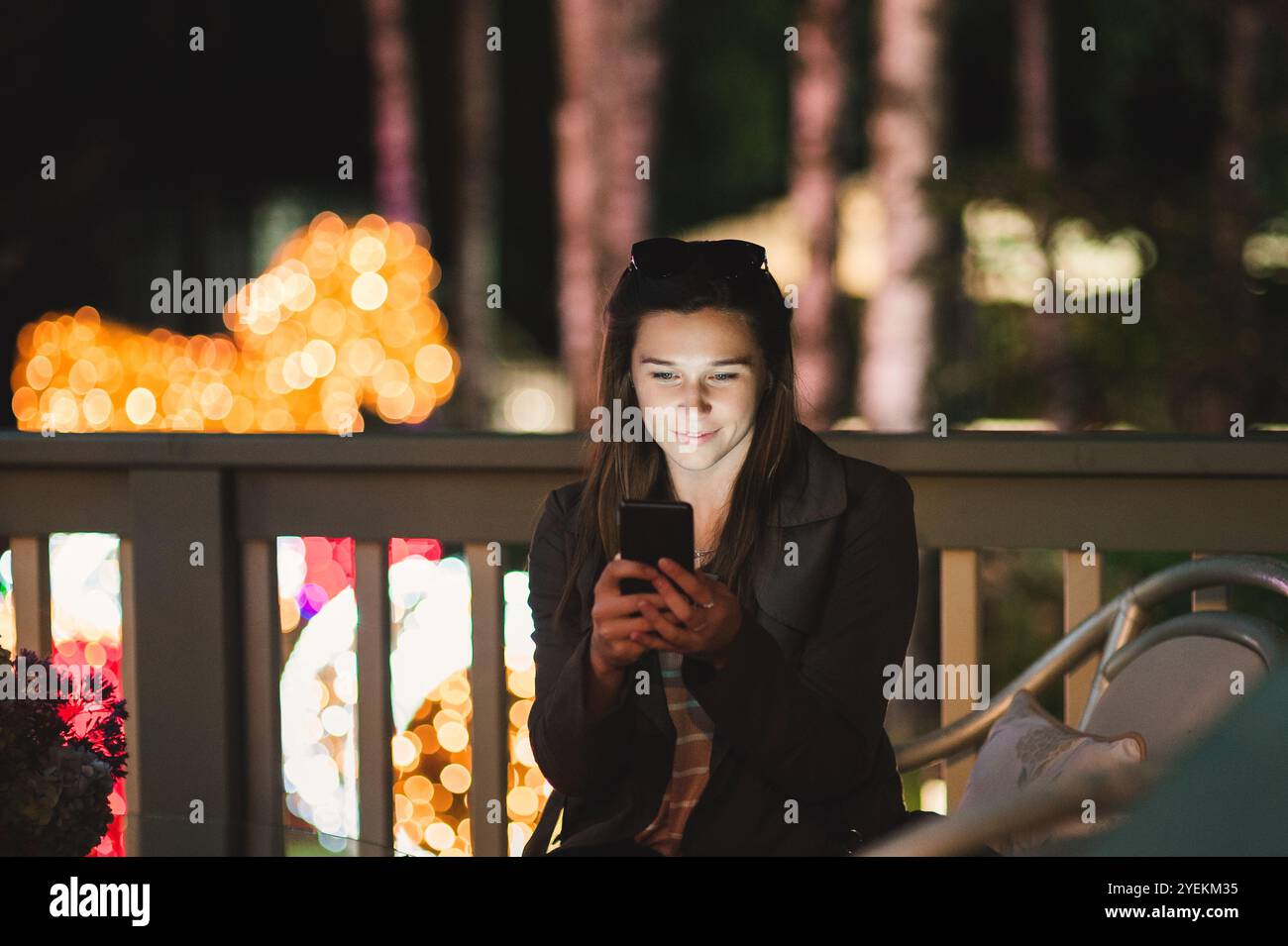 Una giovane donna sorridente che usa uno smartphone di notte. È seduta in giardino con una casa sullo sfondo Foto Stock