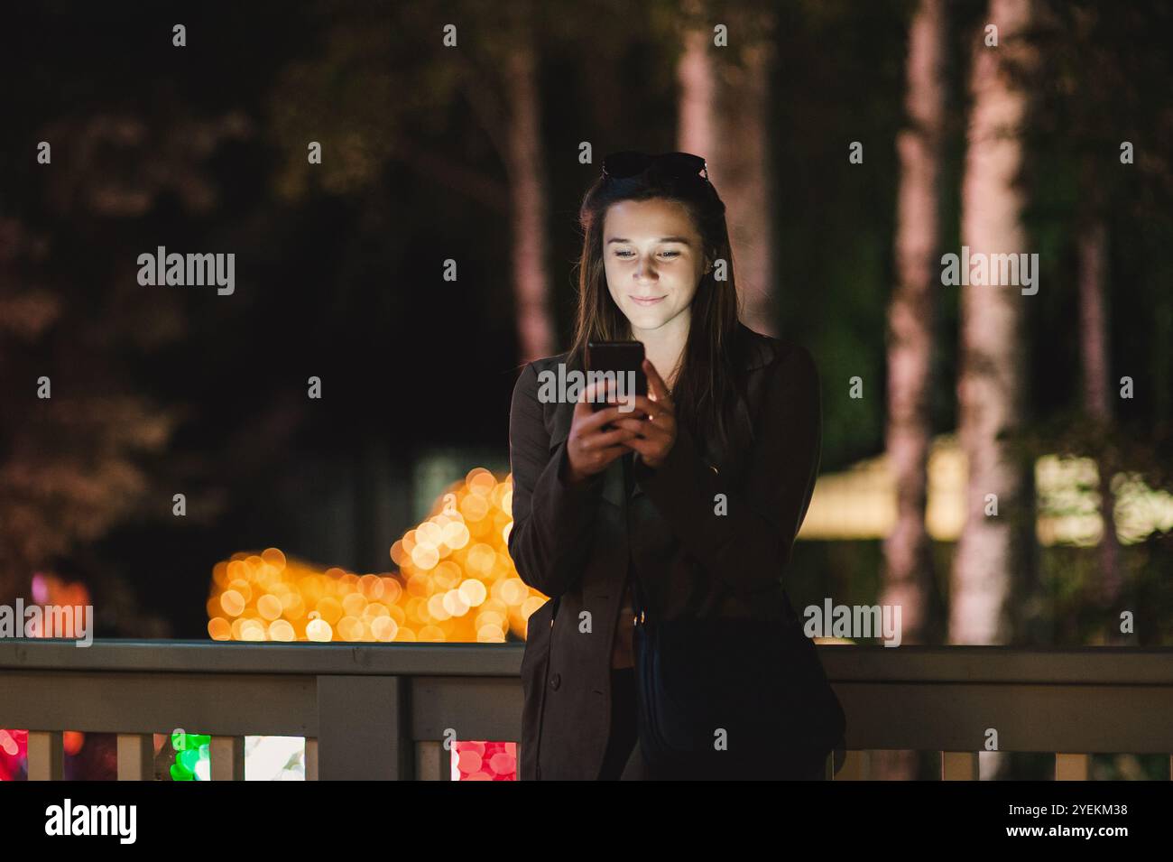 Una giovane donna sorridente che usa uno smartphone di notte. È seduta in giardino con una casa sullo sfondo Foto Stock