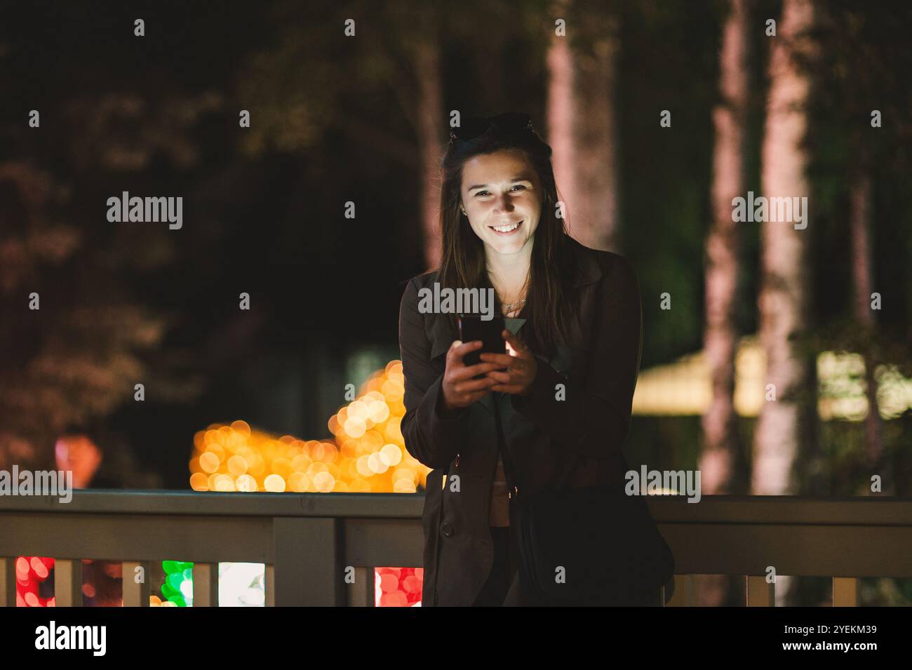 Una giovane donna sorridente che usa uno smartphone di notte. È seduta in giardino con una casa sullo sfondo Foto Stock