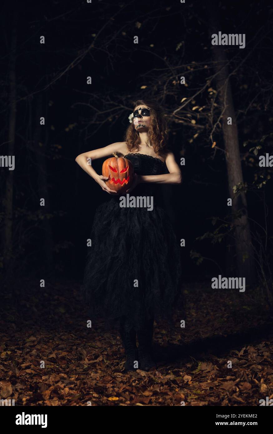 Una strega con un elegante rivestimento per il trucco che tiene in mano una zucca illuminata e una spaventosa Jack-o' lanterna nella foresta durante Halloween Foto Stock