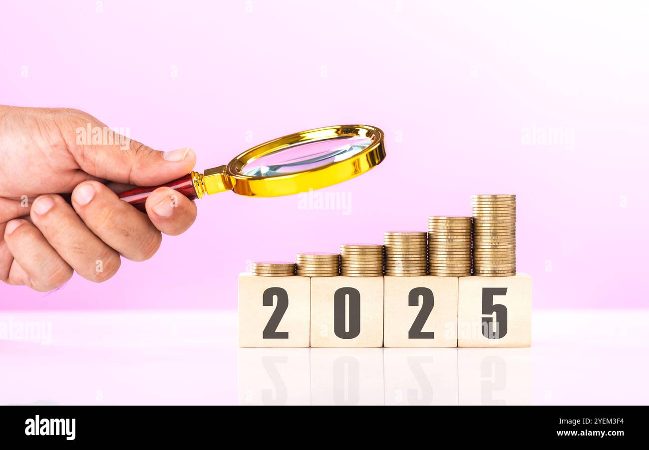 Una pila di monete, un blocco di legno etichettato "2025" e una mano che regge una lente d'ingrandimento. Esame dei piani finanziari e degli investimenti per il prossimo anno Foto Stock