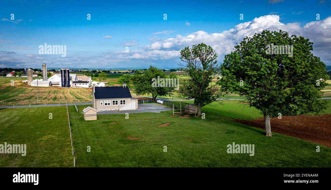 Una casa scolastica Amish a una stanza si trova su un'area di ghiaia, circondata da ampi campi verdi, fienili e strutture lontane sotto un cielo blu brillante. Foto Stock