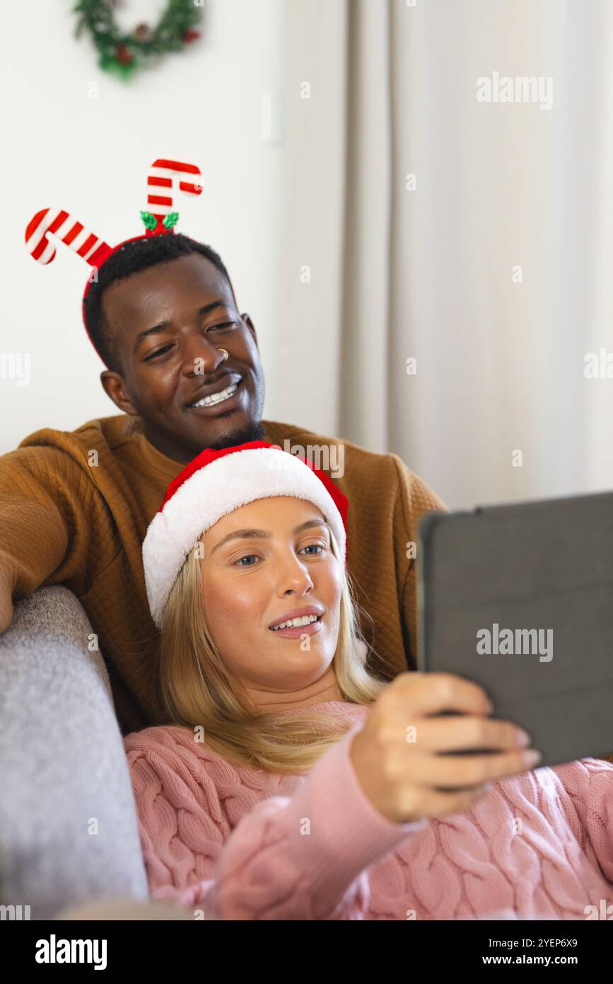 Natale, coppia multirazziale che indossa cappelli festivi e scatta selfie con un tablet a casa. Vacanze, festeggiamenti, tecnologia, felicità, autoritratto, Foto Stock