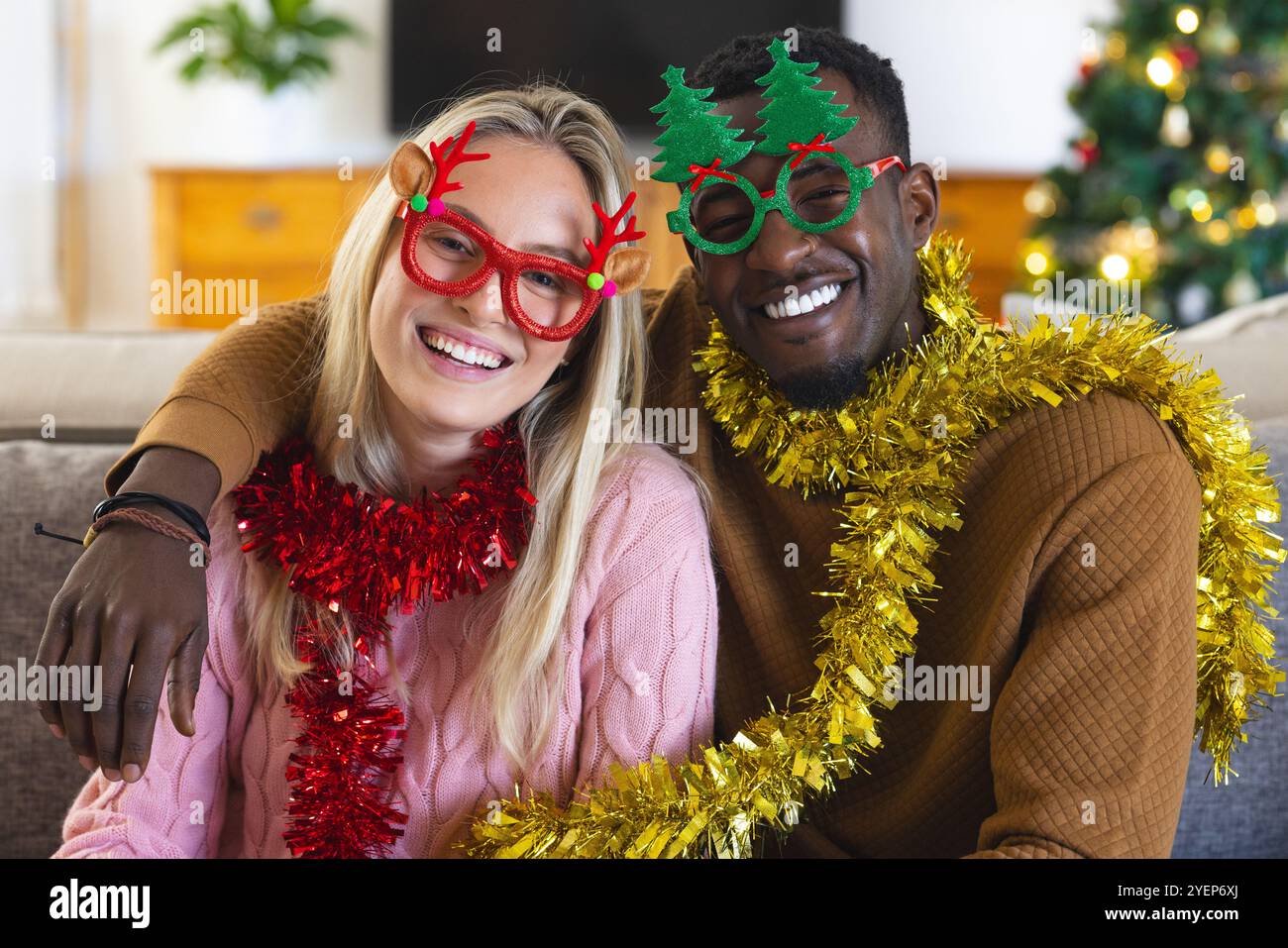 Coppia multirazziale che indossa occhiali e tinsel per festeggiare il Natale a casa. Vacanze, festeggiamenti, felicità, famiglia, diversità, gioioso Foto Stock