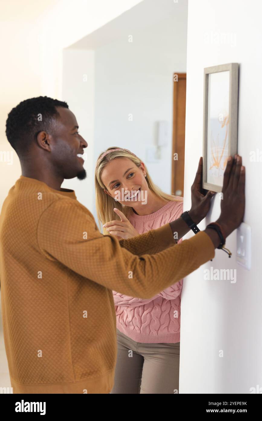 coppia multirazziale con foto a casa, sorridente e divertente. interni, incollaggio, decorazione, felicità, lavoro di squadra, compagnia Foto Stock