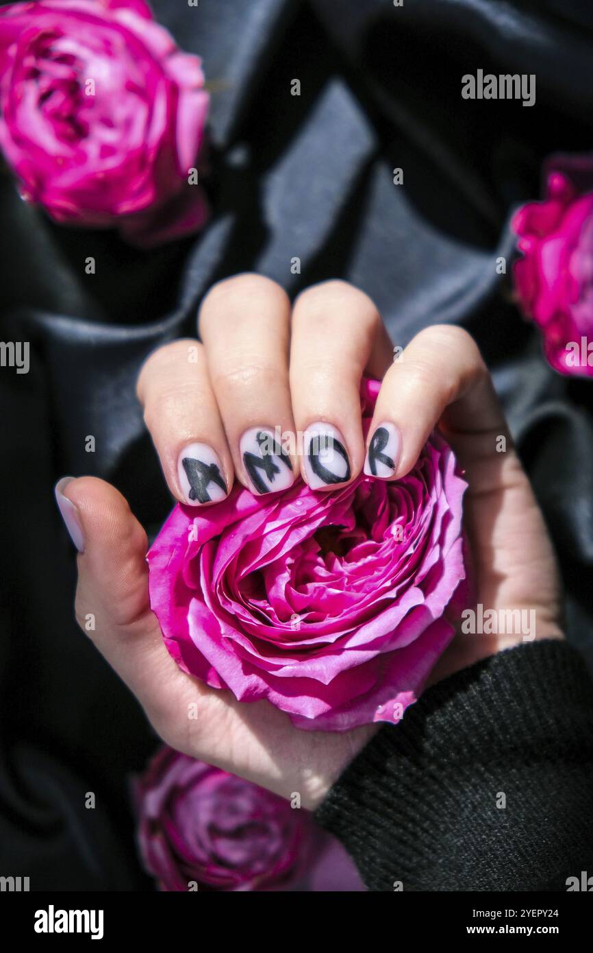 AMOR parola sulle unghie manicure tenere rosa fiore rosa su tessuto di seta nero. Amore, natura piatta minima. Mano femmina. Idea creativa. Cura di sé Foto Stock