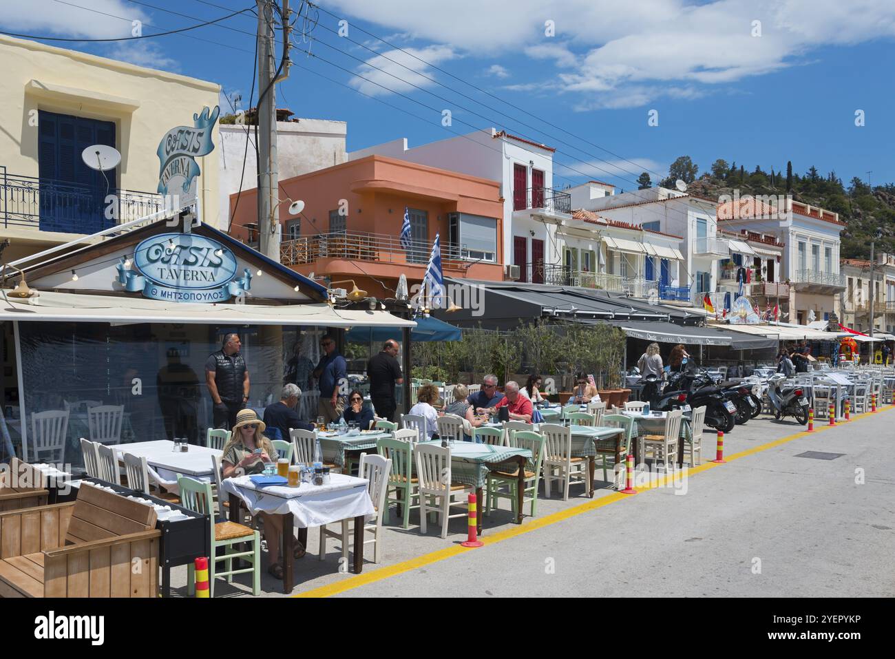 Invitanti ristoranti e taverne fiancheggiano la strada di una città greca, gli ospiti godono dell'atmosfera soleggiata, della passeggiata, di Poros, dell'isola di Poros e delle isole Saroniche Foto Stock