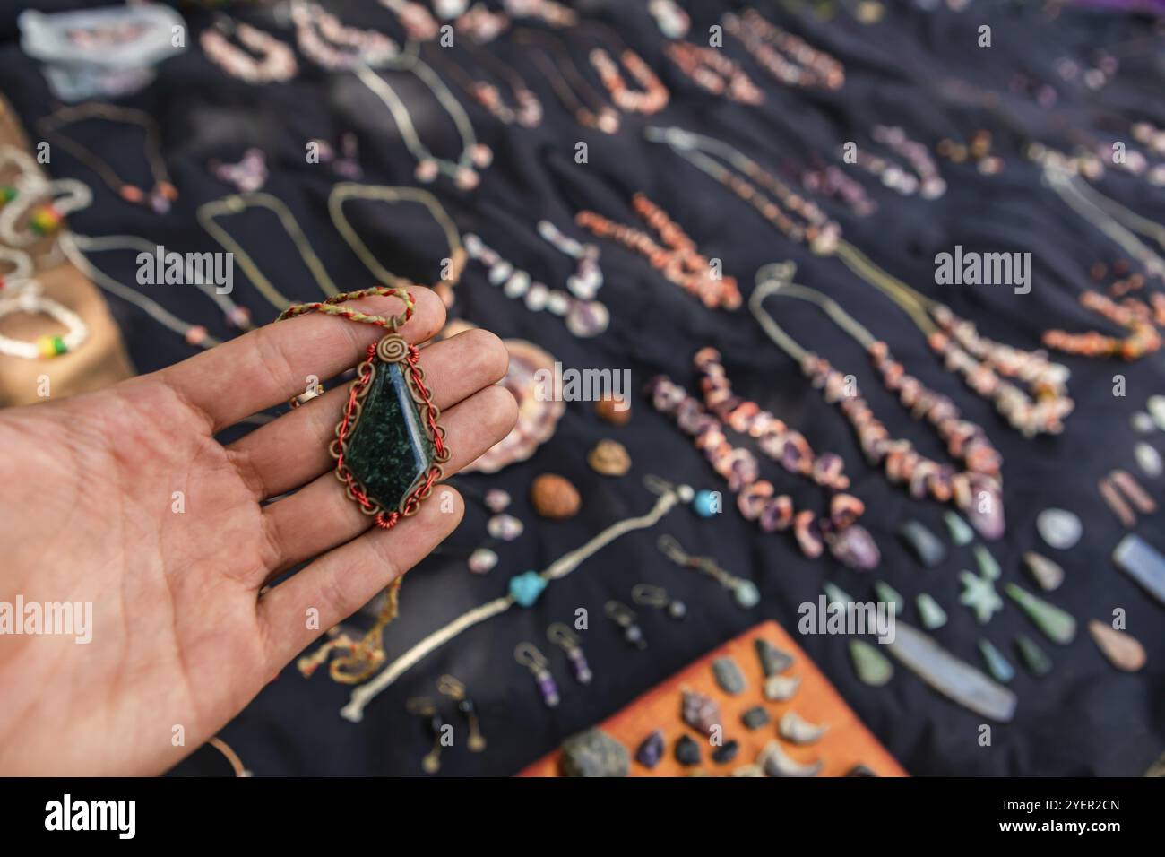 Mano di un uomo che tiene in mano un ciondolo in pietra verde scuro ad un banco di gioielli fatto a mano. Commerciante agricolo, molti altri gioielli e pietre sullo sfondo Foto Stock