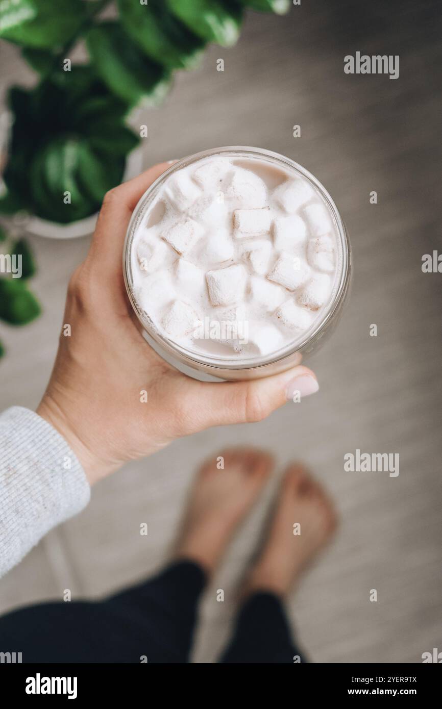 Giovane donna millenaria che tiene una tazza di cacao con marshmallows a casa. Bevanda calda al cioccolato. Pianta su sfondo. Ti sentirai come a casa Foto Stock