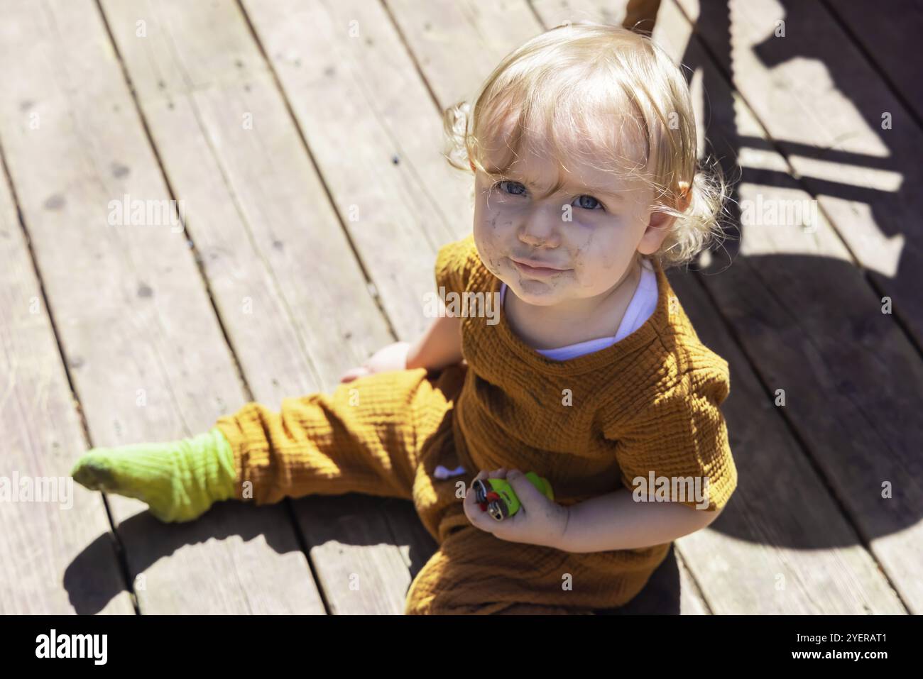 Vista ad angolo alto di un bambino di tre anni malvagio seduto su un ponte di legno che tiene gli accendini con la faccia sporca dopo aver giocato in cortile Foto Stock