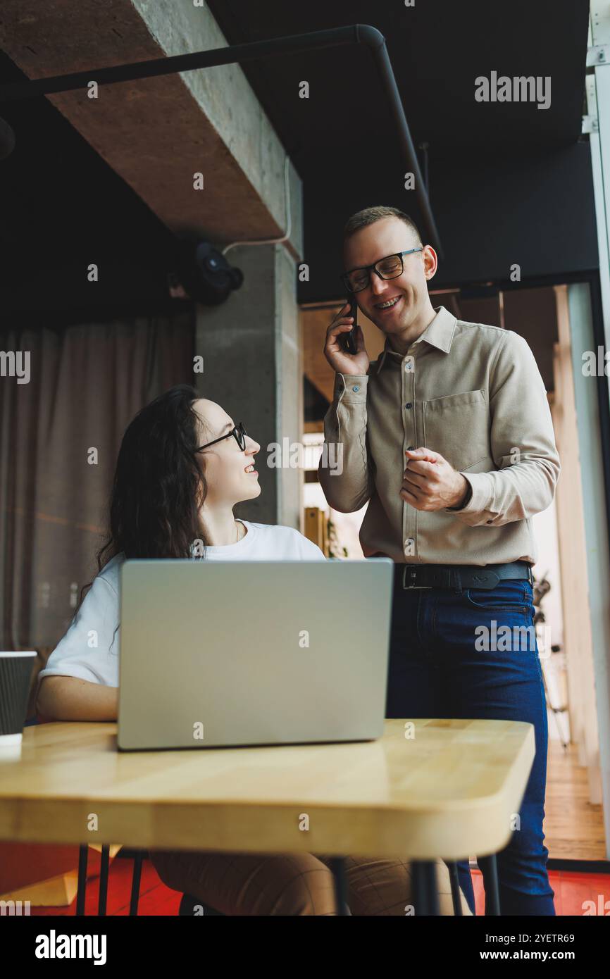 Un giovane uomo soddisfatto parla con una collega e si rallegra del lavoro svolto. I colleghi lavorano al computer portatile in ufficio. Lavorare in un ufficio moderno sp Foto Stock