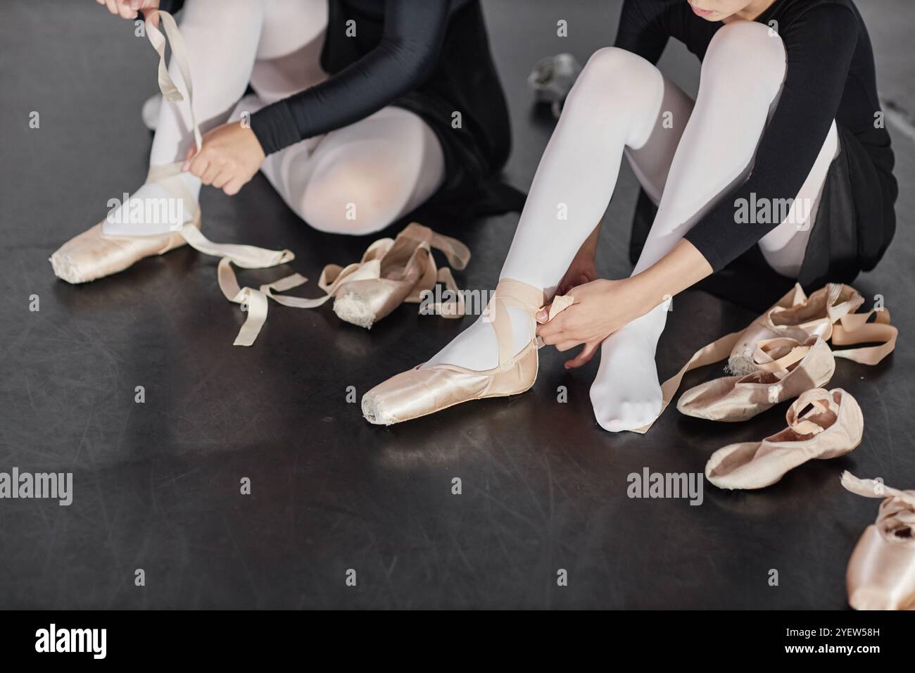 Foto ritagliata di una giovane ragazza che lega scarpe da balletto in raso con nastri in preparazione per la lezione di danza mentre si siede sul pavimento in studio, spazio copia Foto Stock