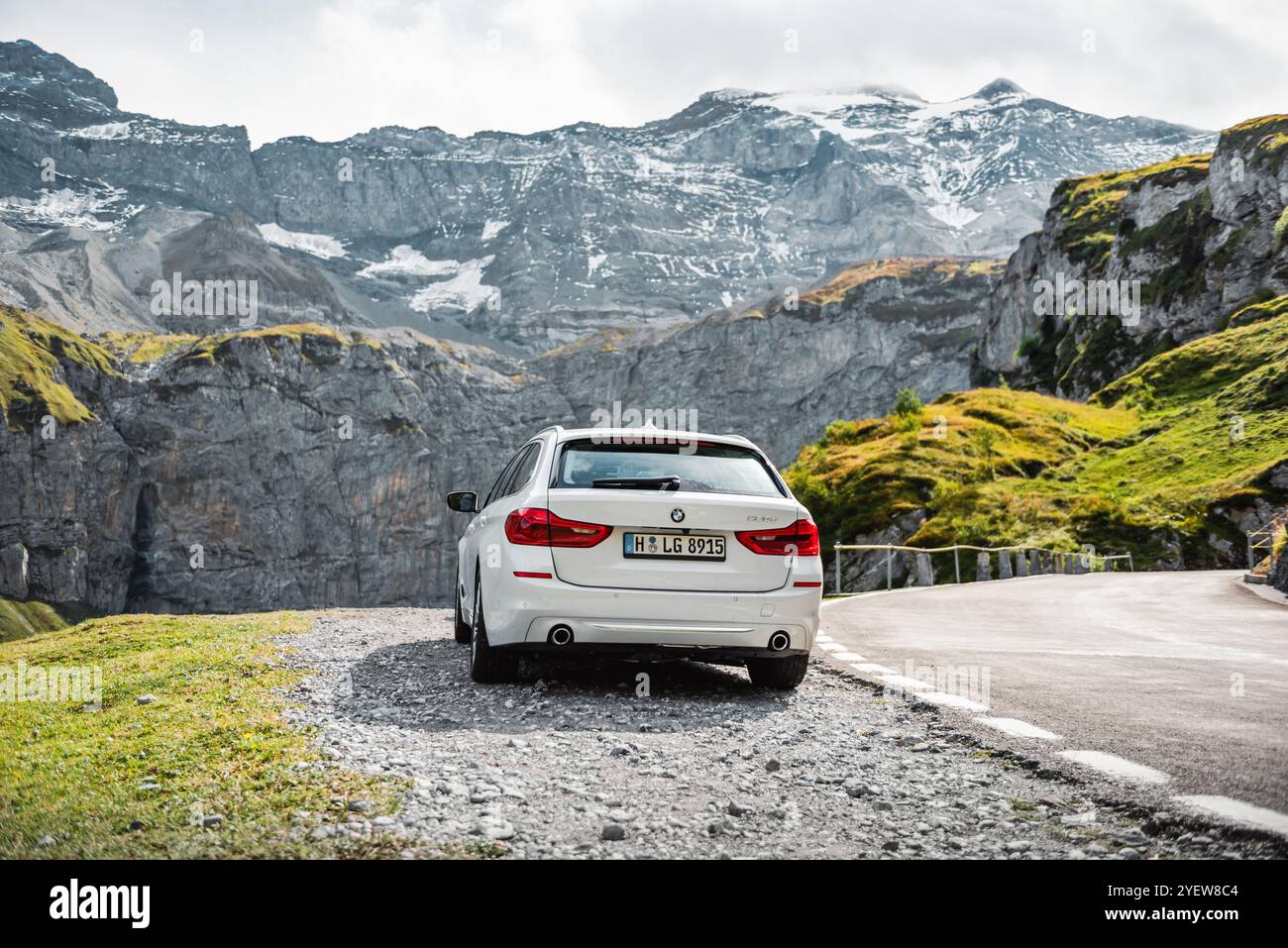BMW serie 5 bianca parcheggiata su un lato di una strada tortuosa nelle alpi svizzere. Vista posteriore dell'auto tedesca, della scogliera e delle cime innevate sul retro. Foto Stock