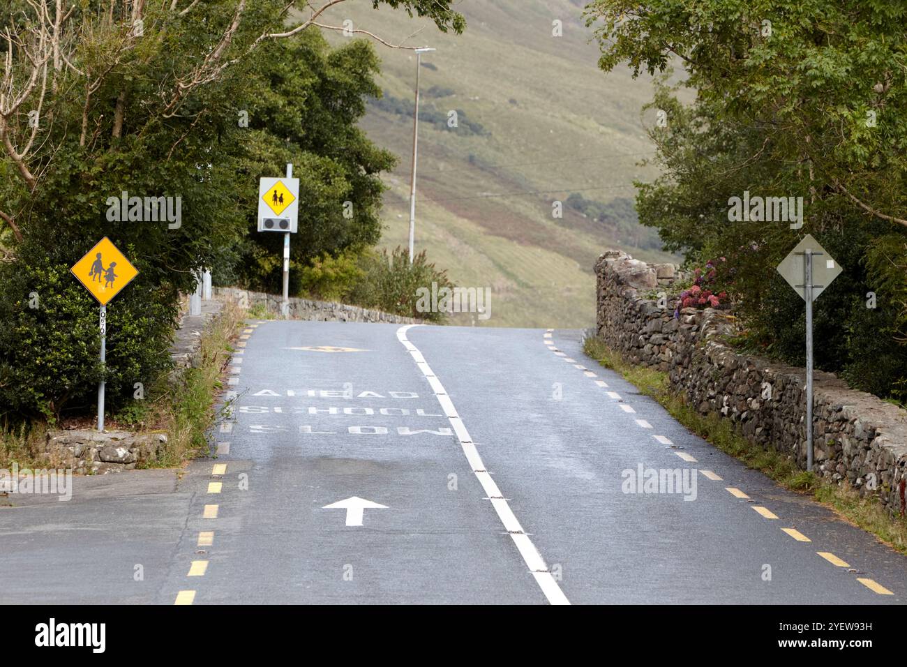 piccola strada rurale stretta su una collina e intorno a una curva che si avvicina a scuola con avanti lento dipinto sulla strada con segnali di avvertimento, contea galway, repubblica Foto Stock