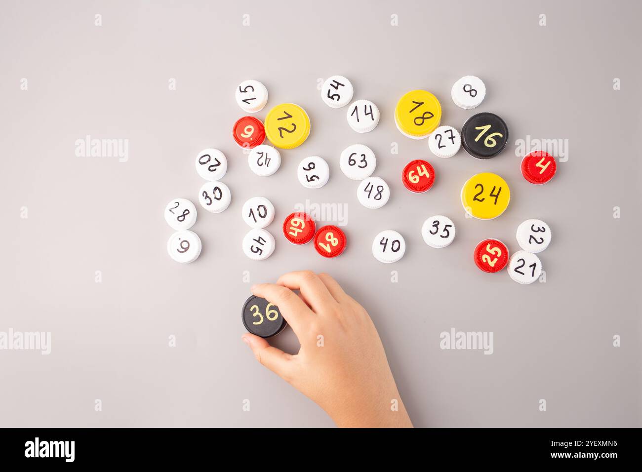 Mani dei bambini che sistemano tappi per bottiglie in plastica con numeri scritti a mano e dati moltiplicati su sfondo grigio. Attività didattica di matematica per ki Foto Stock