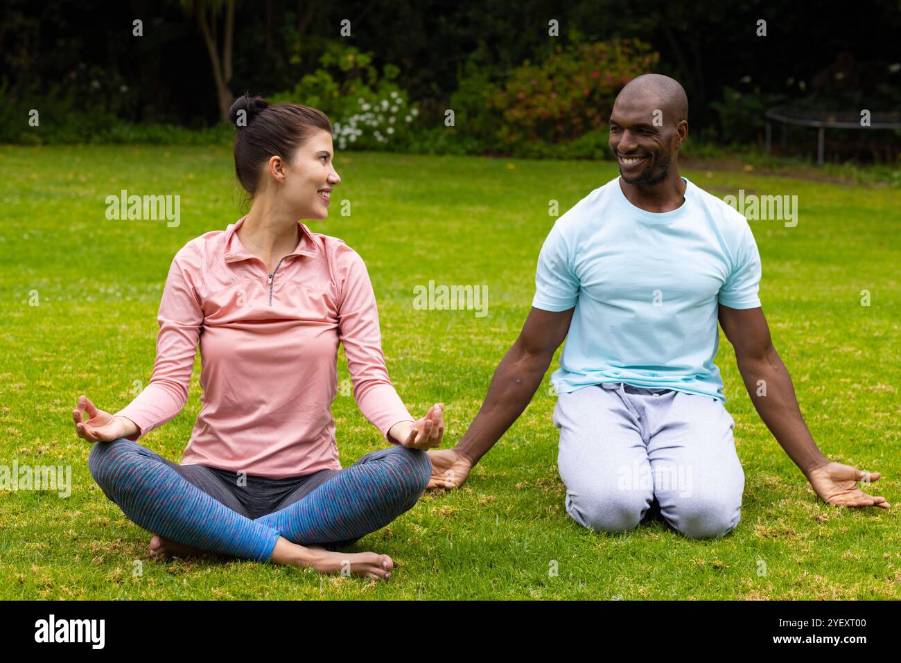 coppia multirazziale che pratica yoga insieme sull'erba, sorridendo e godendo del relax all'aperto Foto Stock