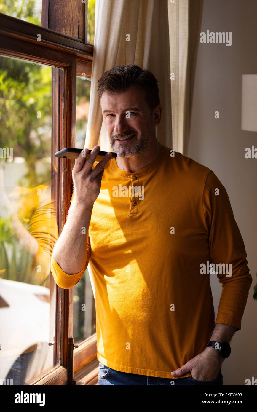Un anziano sorridente che utilizza l'assistente vocale dello smartphone accanto a una finestra soleggiata a casa Foto Stock