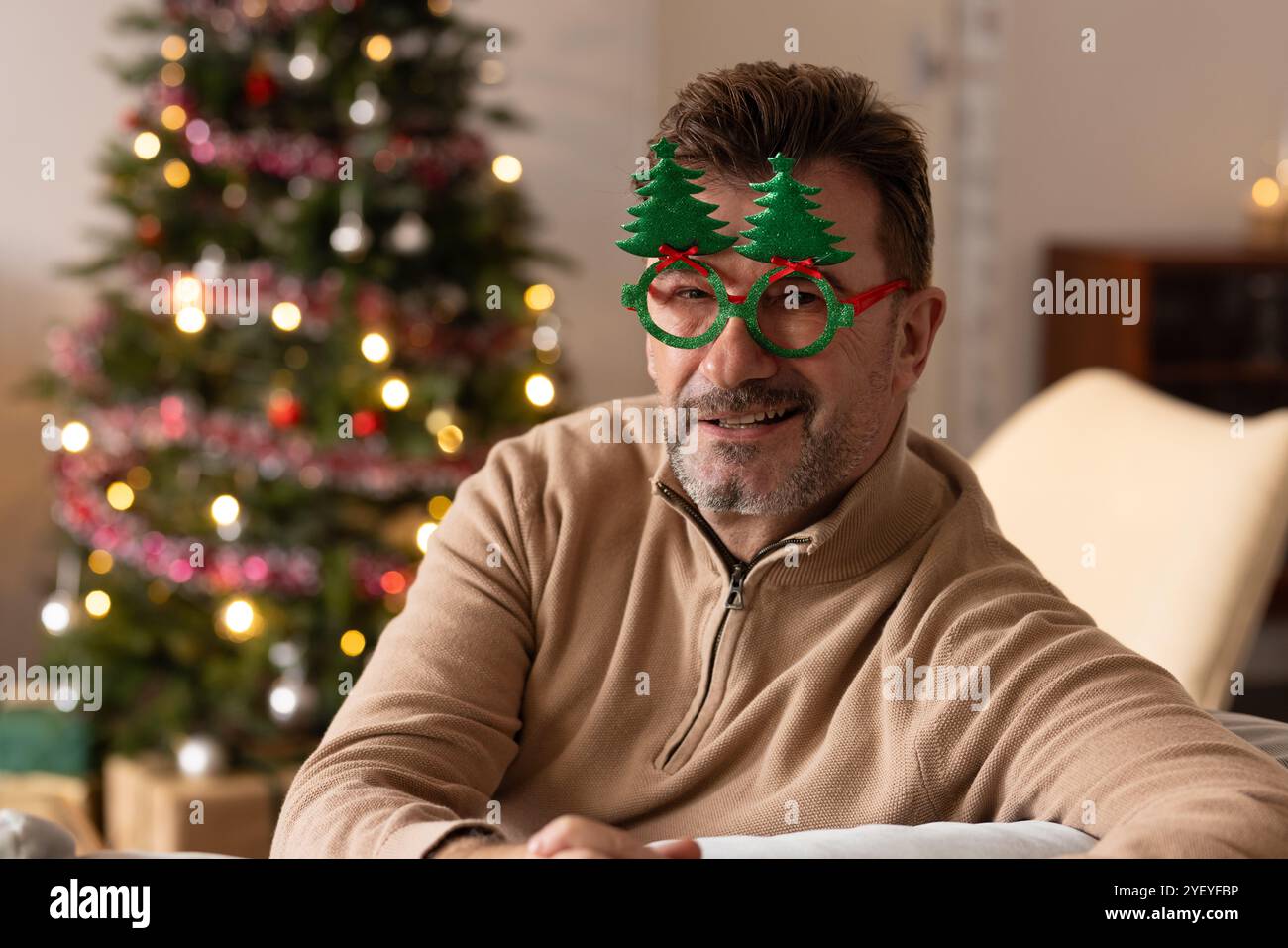 Uomo anziano sorridente che indossa occhiali festivi seduto accanto all'albero di Natale a casa Foto Stock