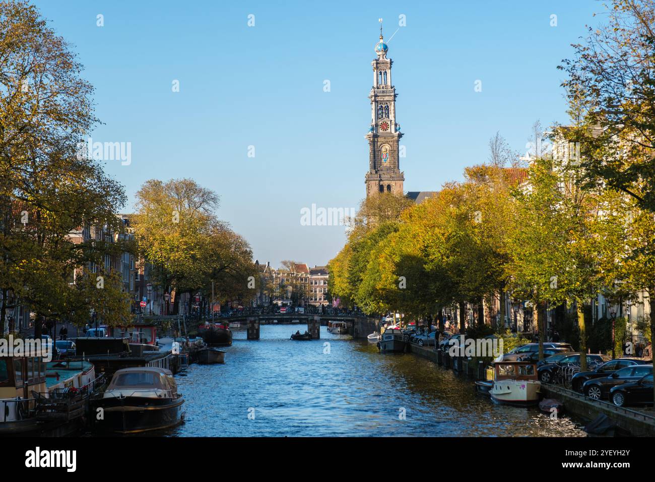 Le foglie d'oro scendono dolcemente dagli alberi che costeggiano i tranquilli canali di Amsterdam. L'iconica torre di Westerkerk si erge alta mentre le barche galleggiano pacificamente, catturando l'essenza di una giornata autunnale Foto Stock