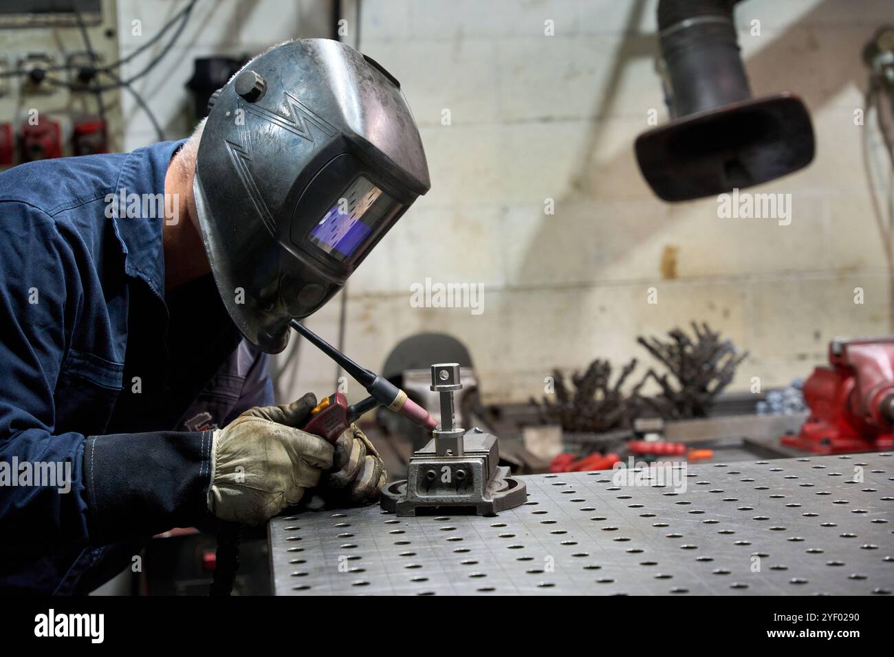 Lavoratore di metalli che indossa una maschera protettiva per la saldatura di una parte meccanica personalizzata in un'officina metallurgica Foto Stock