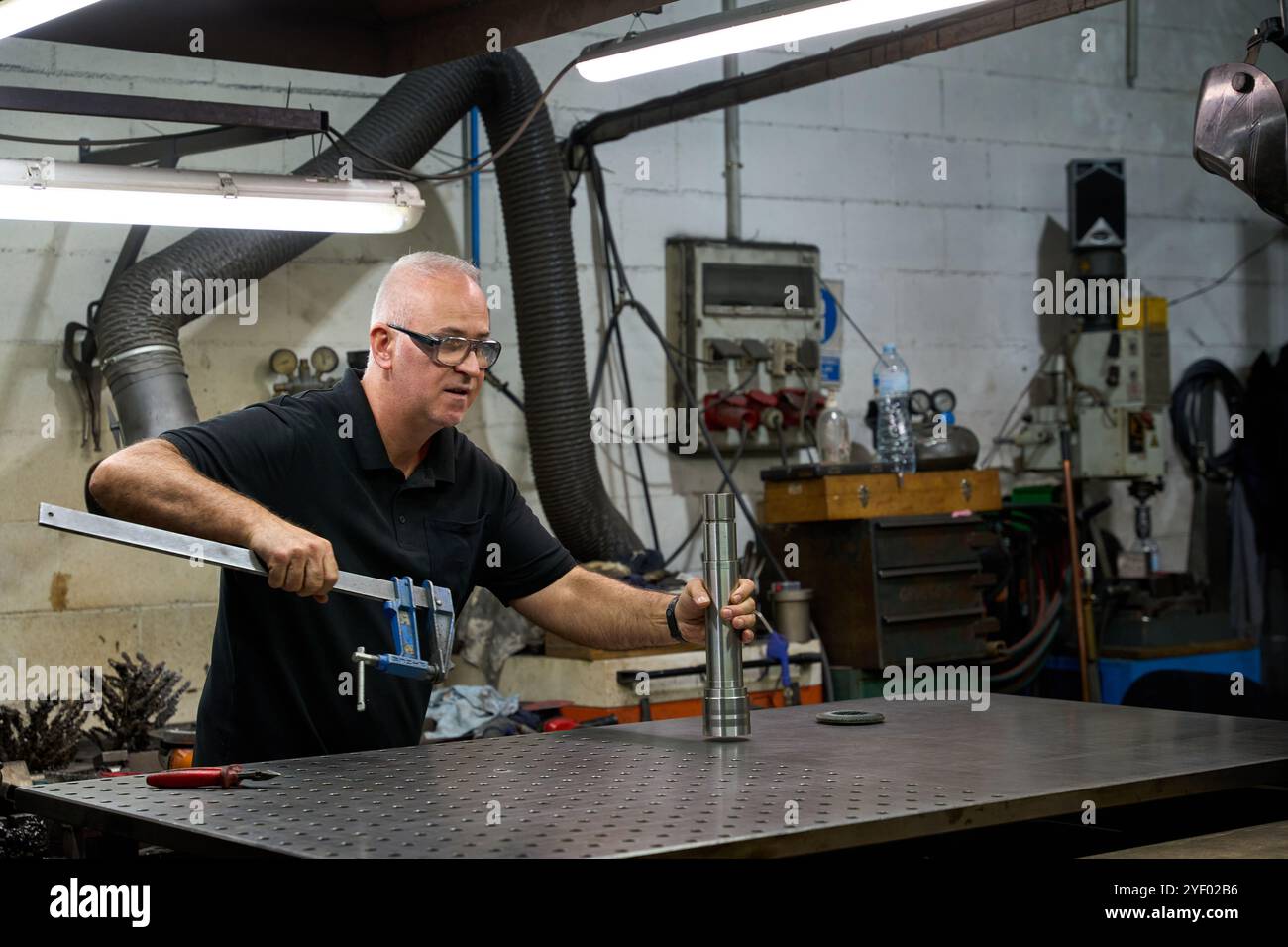 Operaio di metalli che indossa occhiali di sicurezza che tengono un morsetto e che esamina una parte meccanica personalizzata in un'officina metallurgica Foto Stock
