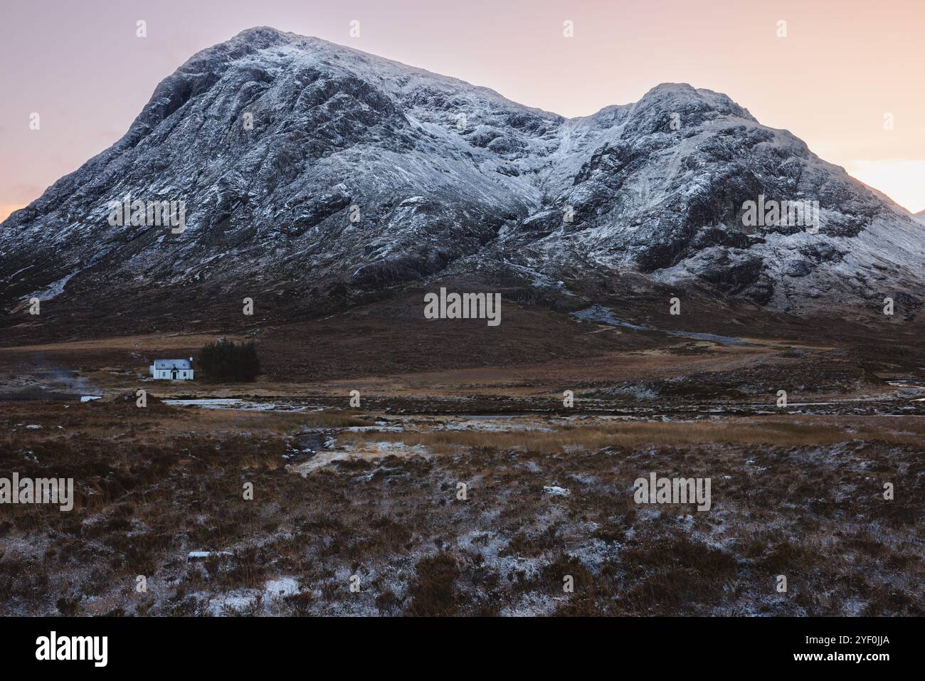 Un remoto cottage si trova ai piedi delle montagne innevate a Glencoe, in Scozia, catturando la bellezza serena delle Highlands scozzesi al tramonto o al sole Foto Stock