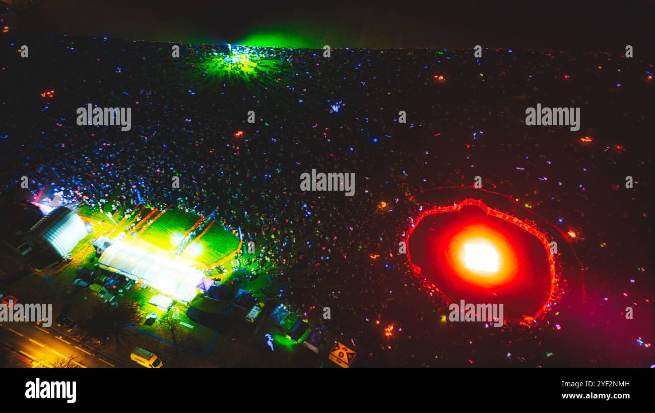 Una vista aerea di una grande folla riunita in un evento notturno, con un palco e un anello di fuoco nel centro. La folla è illuminata da un labbro colorato Foto Stock