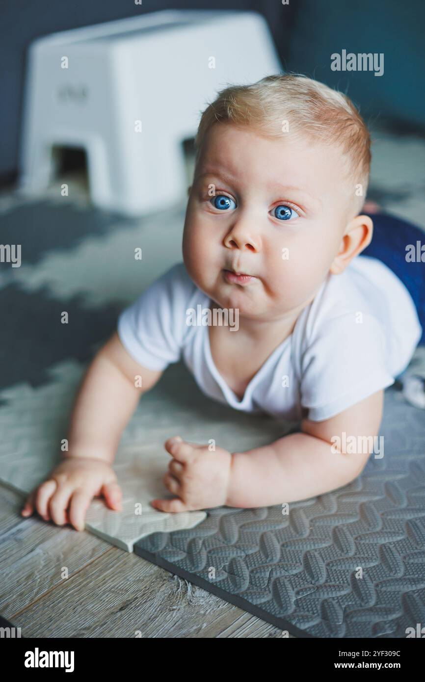 Un bambino dagli occhi blu con un body bianco e pantaloni blu impara a sedersi. Sviluppo personale del bambino. Il bambino è seduto sul pavimento e sorride Foto Stock