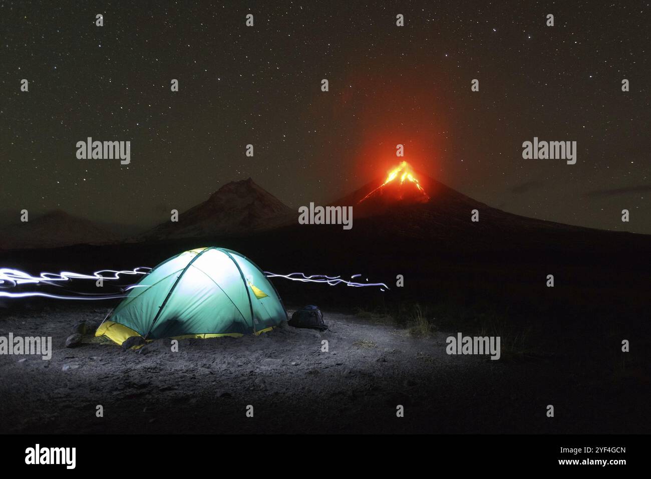 Notte del paesaggio di montagna della penisola di Kamchatka: illuminato campeggio turistico su sfondo eruzione vulcanica, la luce proveniente da proiettori intorno a tenda. La Russia Foto Stock