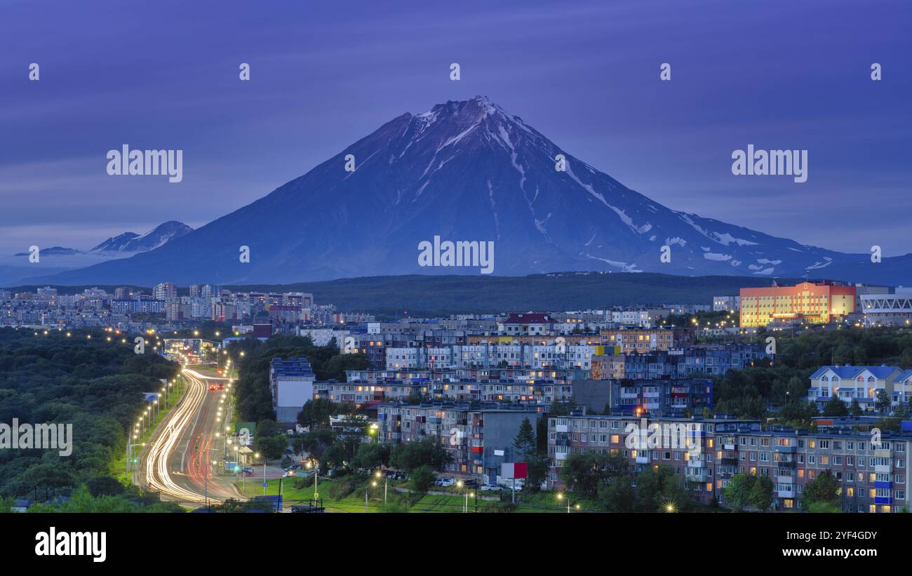 Panorama della città notturna di Petropavlovsk-Kamchatsky sullo sfondo cono del vulcano, sviluppo urbano al crepuscolo, strada della città retroilluminata con auto li Foto Stock